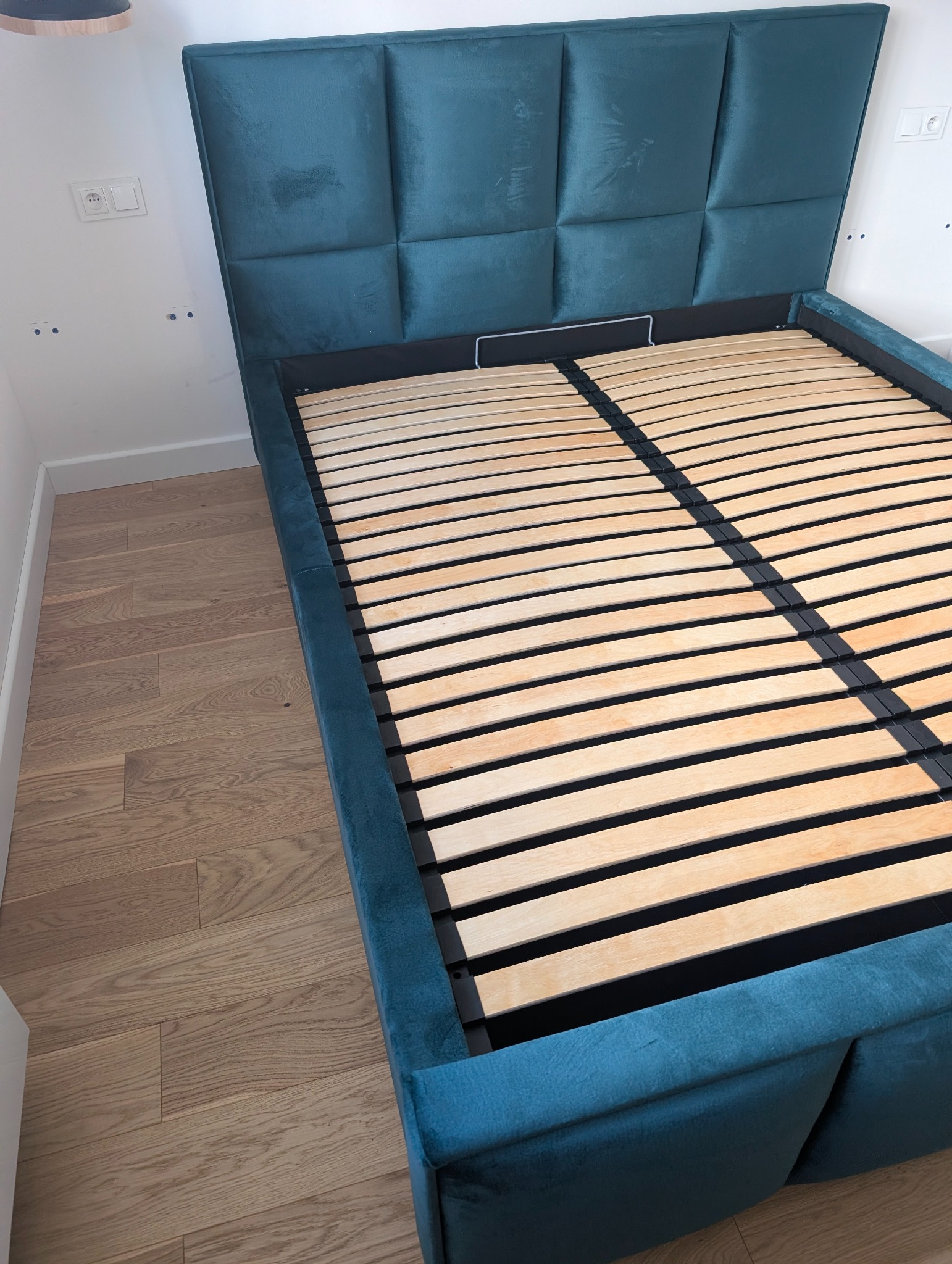 Turkusowe łóżko z tapicerowanym wezgłowiem w kwadraty i drewnianym stelażem, ustawione na parkiecie. Widoczne gniazdka elektryczne na ścianie.
