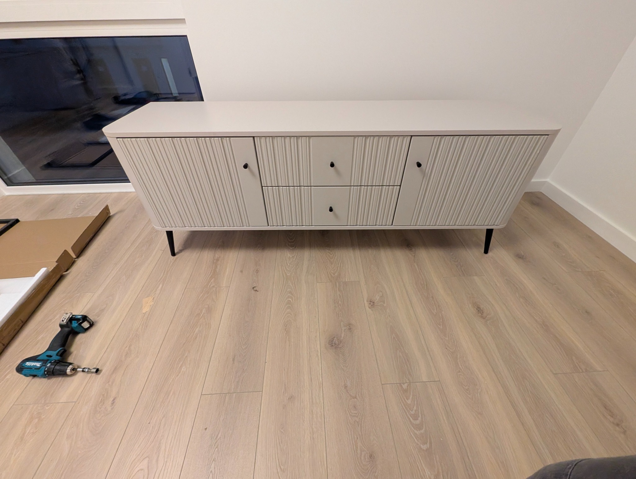 Jasny, gotowy do użytku kredens z ryflowanymi frontami, stojący na drewnianej podłodze. Obok leży wkrętarka i karton po meblach. Widok z góry, naturalne światło.
