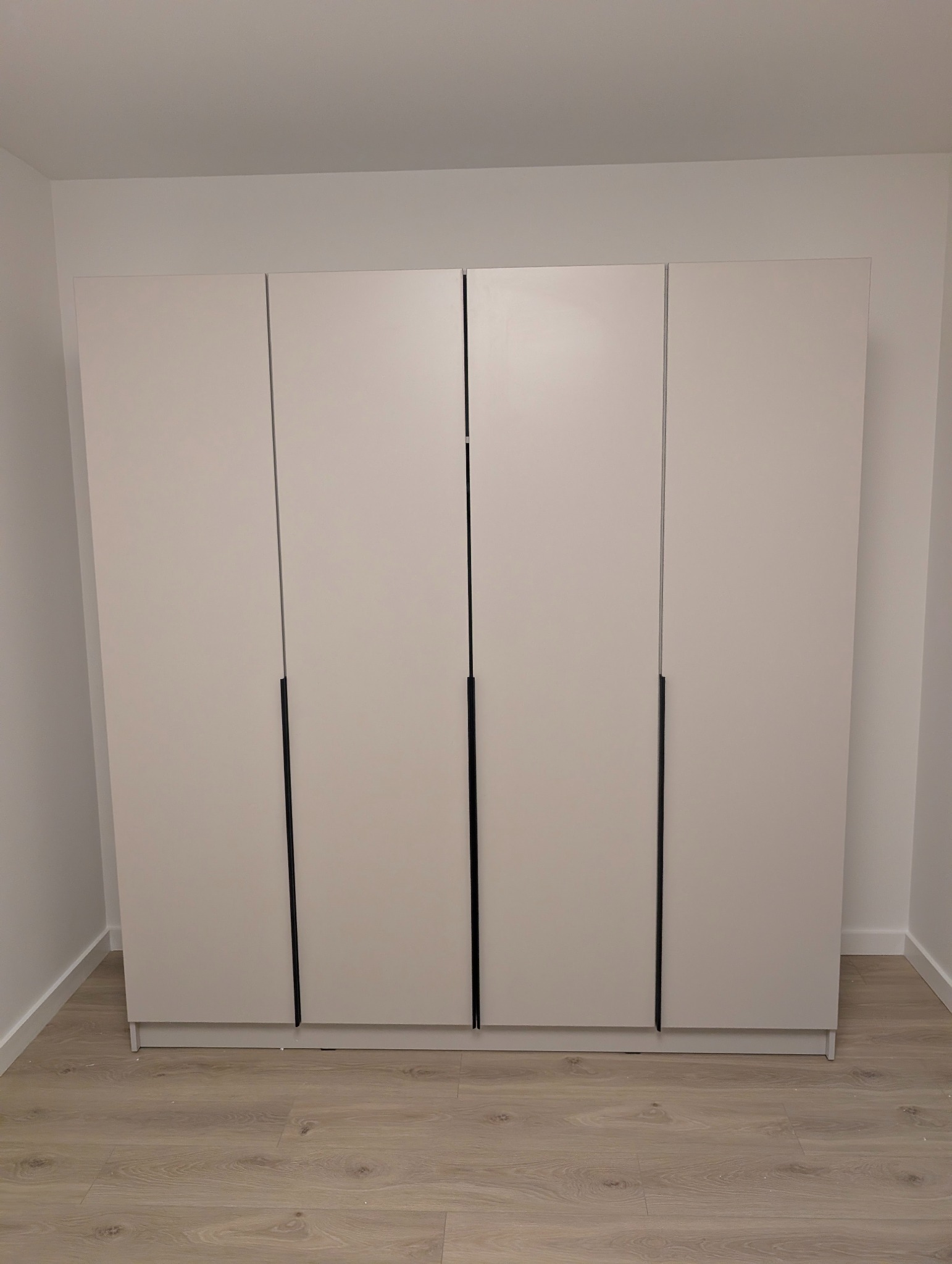 Czterodrzwiowa, biała szafa z czarnymi uchwytami, ustawiona na podłodze z paneli w jasnym odcieniu drewna, w rogu pomieszczenia. Minimalistyczny design.