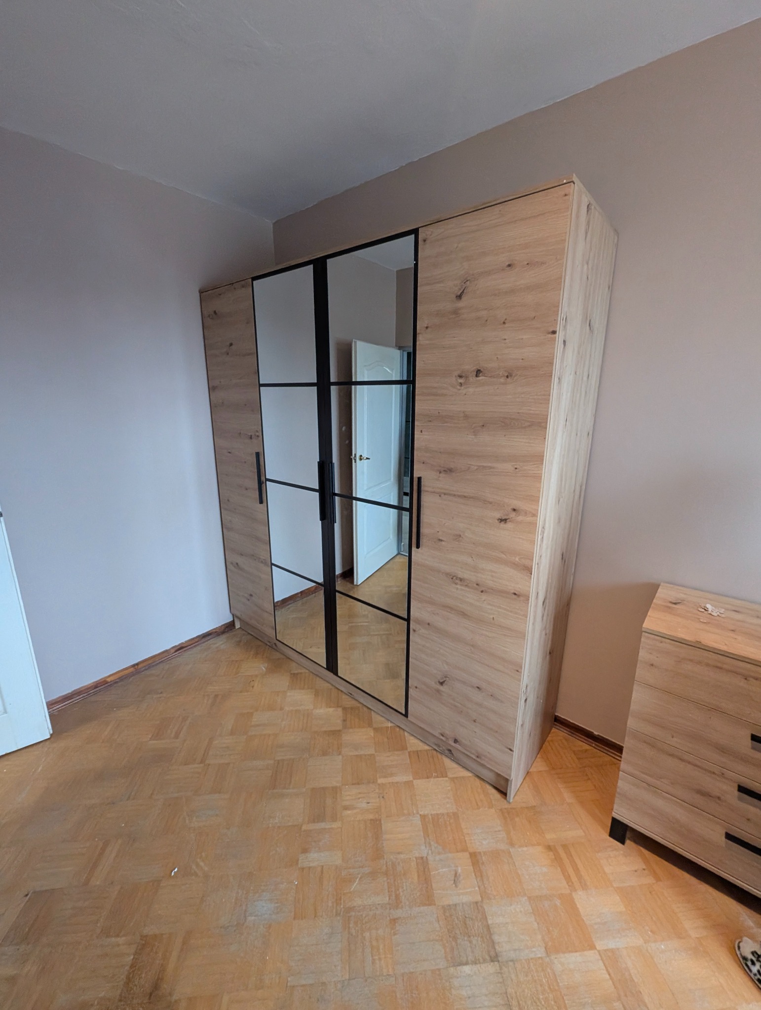 Szafa z lustrami i drewnianymi panelami, czarne uchwyty, widok na wnętrze pokoju przez lustra, parkiet na podłodze, komoda obok szafy.