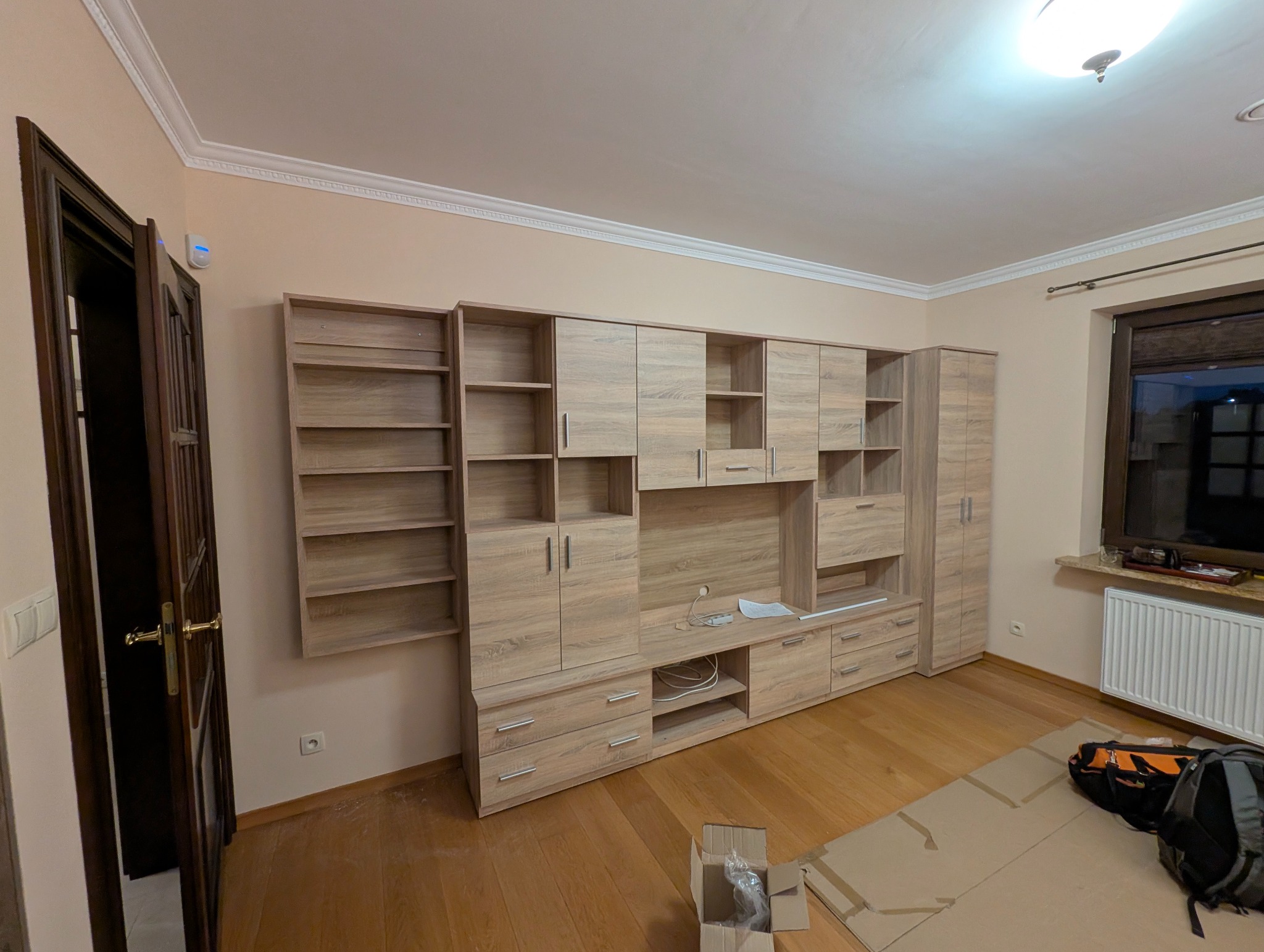 Zmontowana, duża meblościanka w odcieniach drewna, z otwartymi półkami i szafkami, na drewnianej podłodze. Widoczna otwarta, ciemna stolarka drzwiowa i okno.