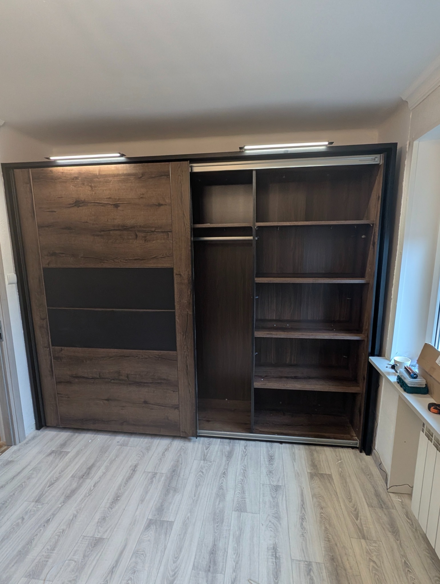 Szafa przesuwna z drewnianymi drzwiami i czarnymi wstawkami, częściowo otwarta, ukazująca półki. Widoczne narzędzia na parapecie. Podłoga laminowana.