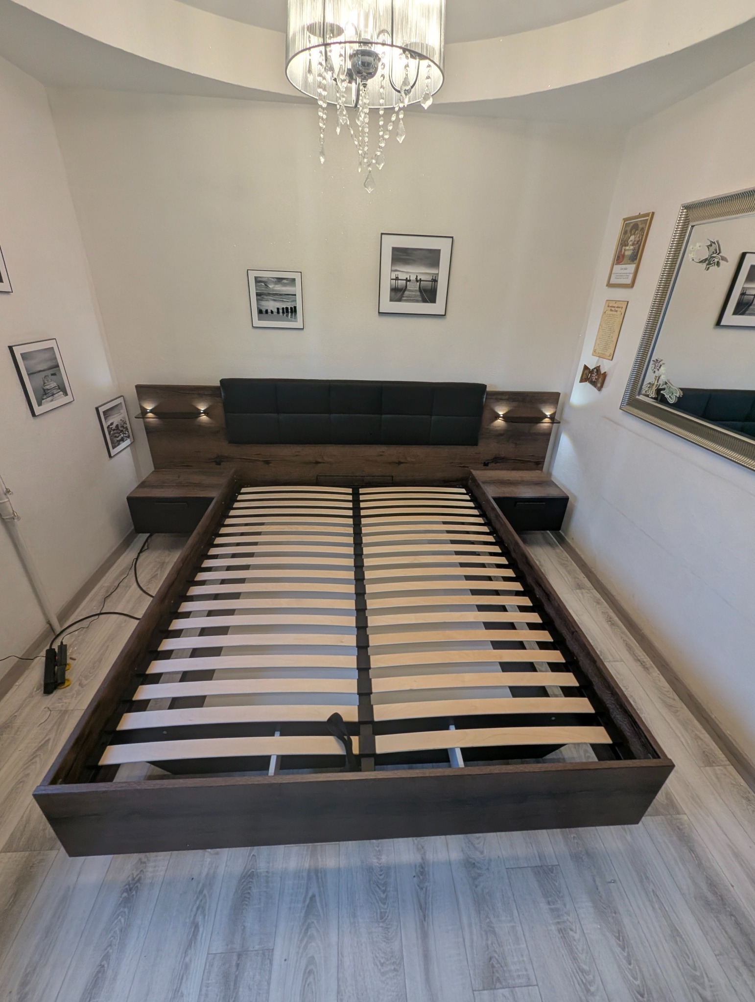 Częściowo zmontowane łóżko z drewnianą ramą i stelażem listwowym, zintegrowane szafki nocne i oświetlenie, w jasnym wnętrzu z dekoracjami ściennymi.