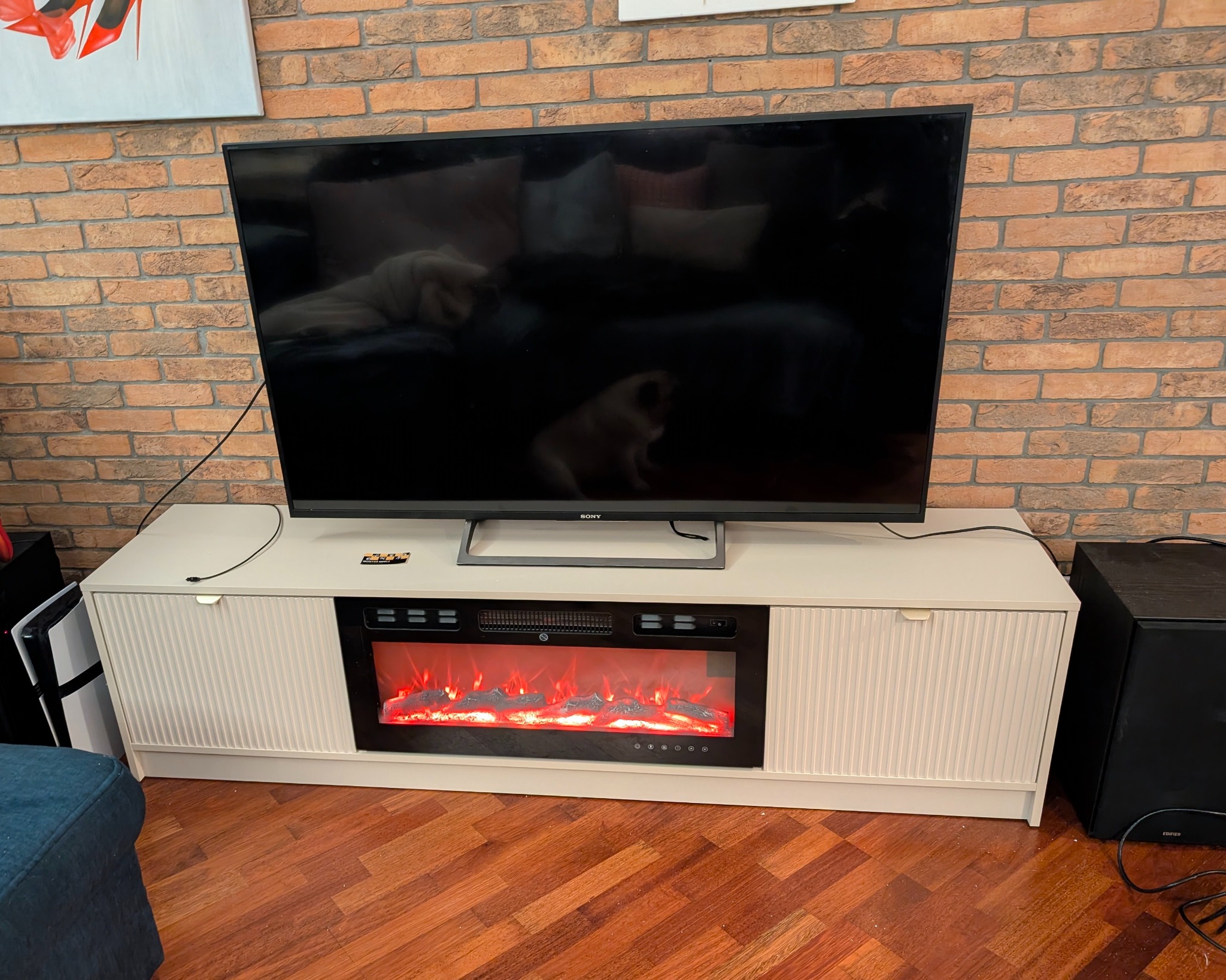 Biała szafka RTV z elektrycznym kominkiem pod telewizorem Sony, na tle ceglanej ściany. Widoczny fragment podłogi i konsola do gier.