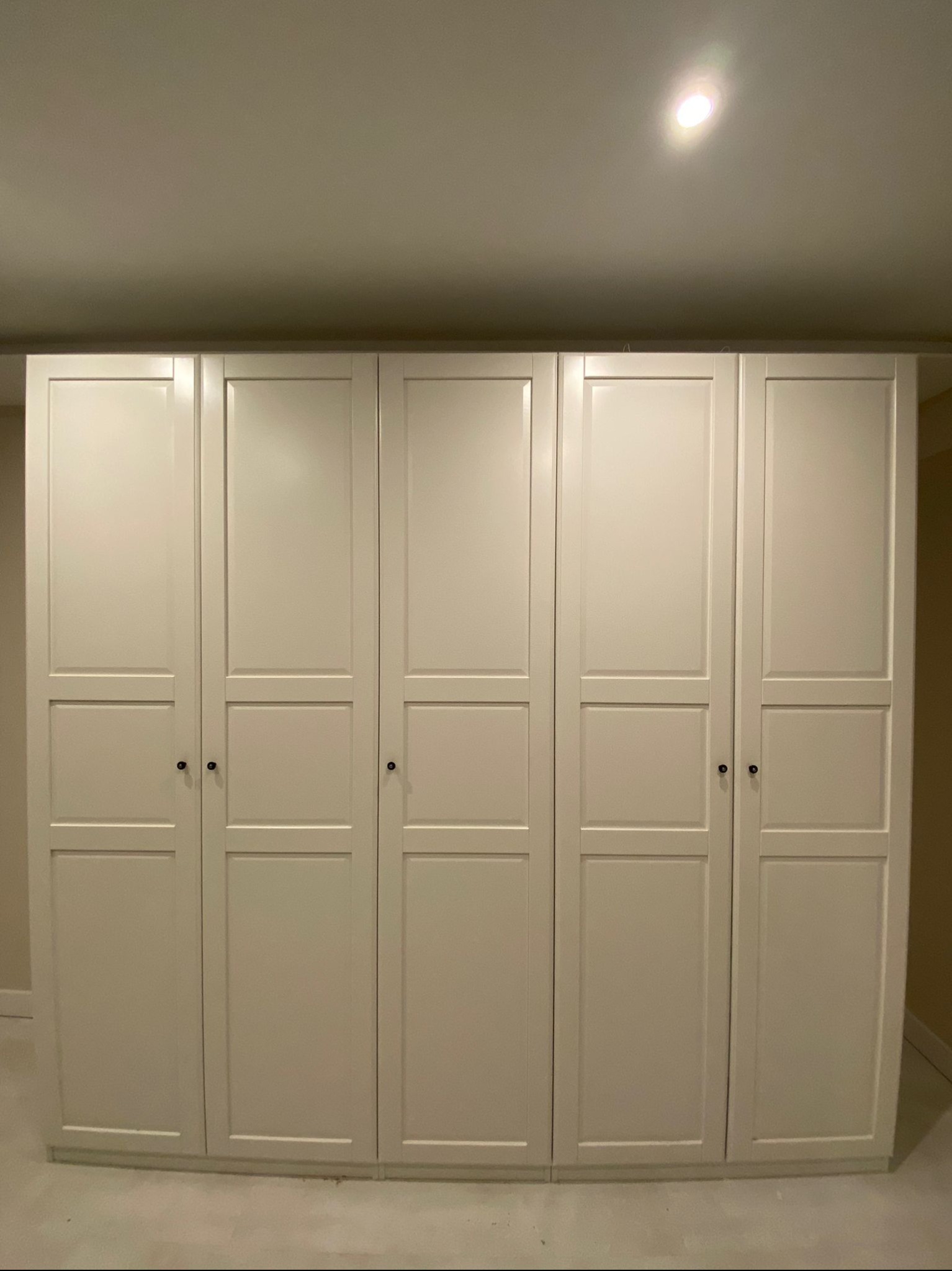 Zamontowana, biała szafa z sześcioma drzwiami, z czarnymi uchwytami, oświetlona punktowym światłem sufitowym. Widok frontalny, równomierne oświetlenie.