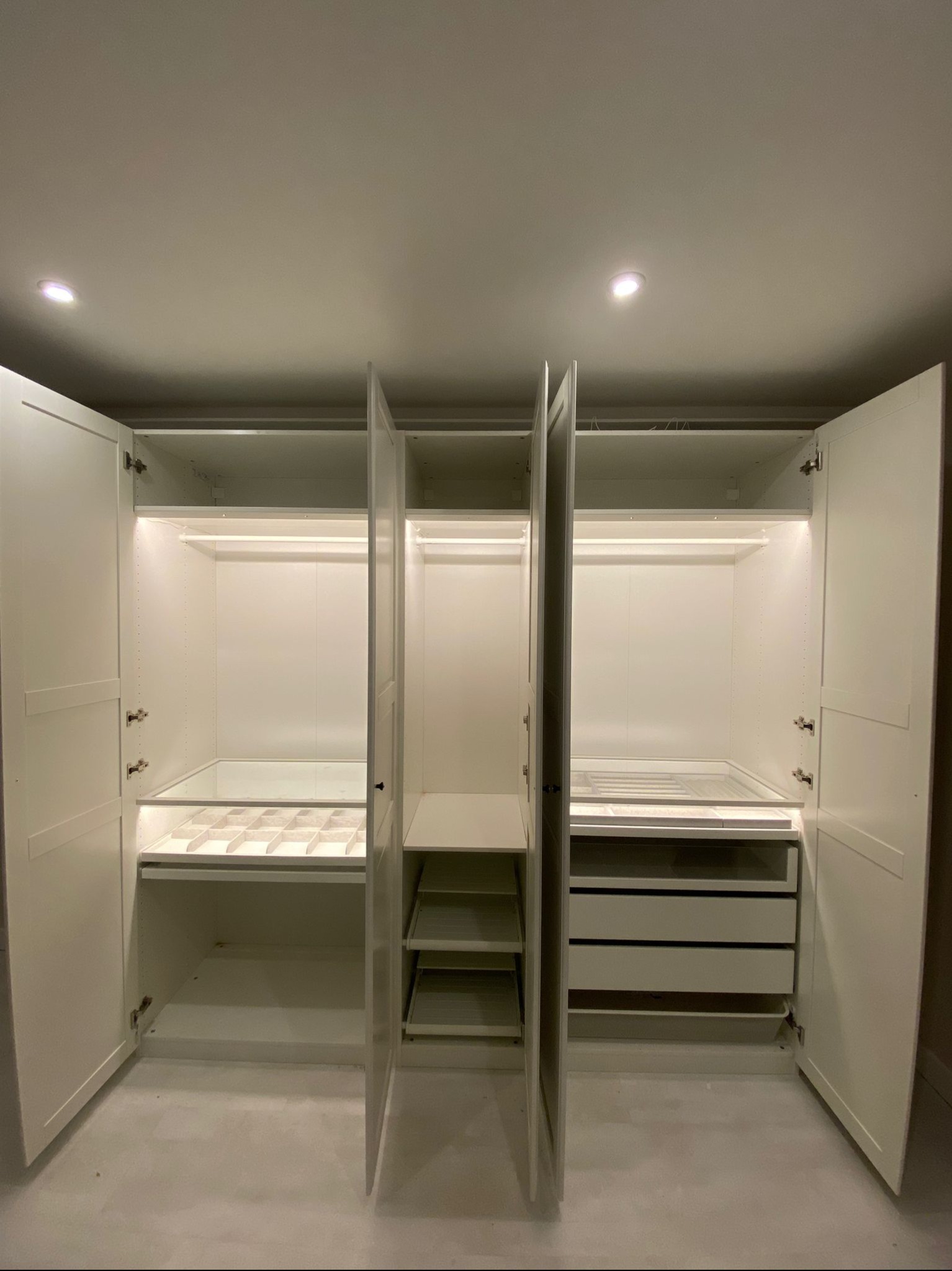 Biała, otwarta szafa wnękowa z oświetleniem LED, widoczne półki, szuflady i drążki na ubrania. Minimalistyczny design, jasne wnętrze.