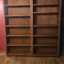 Dwie brązowe, drewniane szafy bez drzwi, z półkami, stojące obok siebie na drewnopodobnej podłodze. Tło stanowią ściany w kolorze fuksji i ciemnej zieleni.