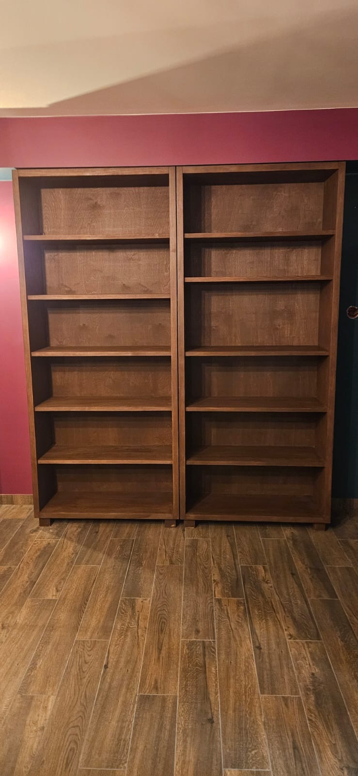 Dwie brązowe, drewniane szafy bez drzwi, z półkami, stojące obok siebie na drewnopodobnej podłodze. Tło stanowią ściany w kolorze fuksji i ciemnej zieleni.