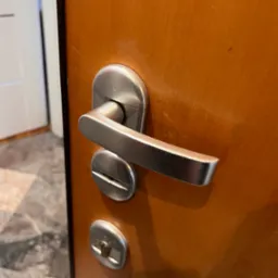 Brązowe drzwi z zamontowaną, srebrną klamką i zamkami. Widoczny fragment podłogi i drzwi w tle. Ujęcie z bliska, skupione na detalach.