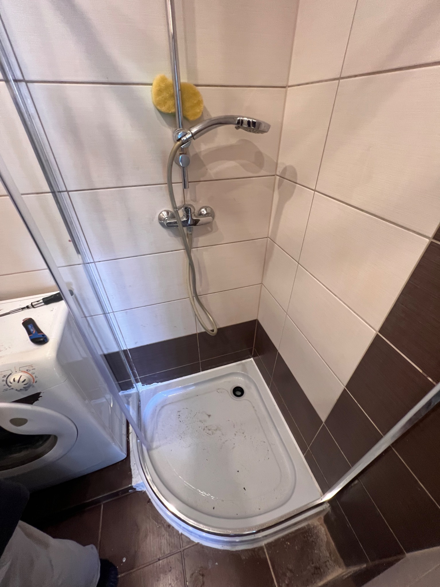 Narożna kabina prysznicowa z chromowaną armaturą i żółtą gąbką, widoczna pralka i narzędzia. Ujęcie z góry, ukazujące detale wykończenia i stan kabiny.