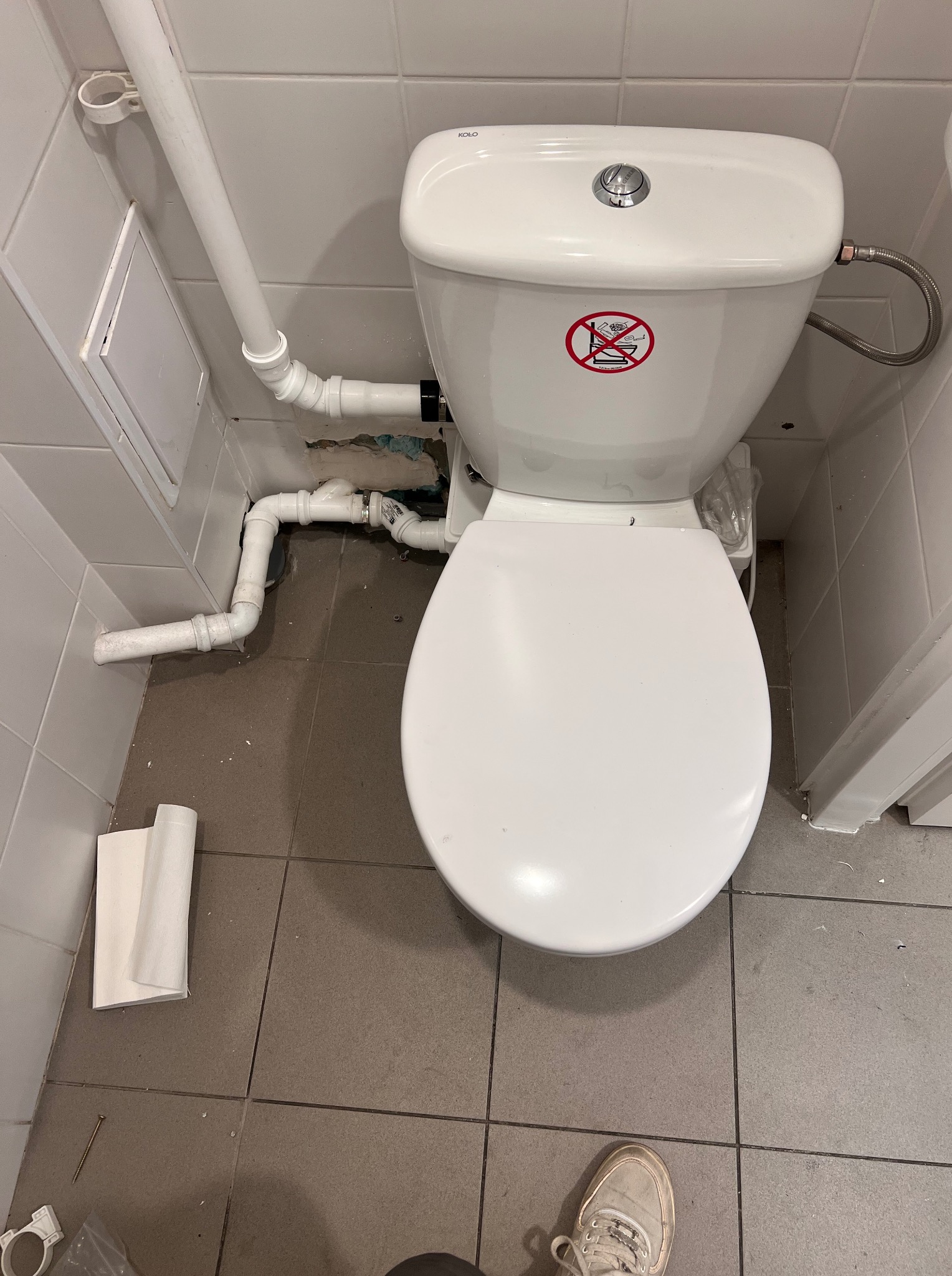 Biała toaleta w łazience z widocznymi rurami i elementami instalacji. Na sedesie naklejka z zakazem wrzucania przedmiotów. Widoczne niedociągnięcia w wykończeniu ściany.