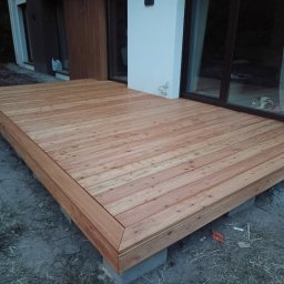 Łukasz Dziklinski - Nowoczesny taras drewniany z jasnego drewna, wykończony listwami, na betonowych bloczkach, przylegający do budynku z dużym oknem przesuwnym.