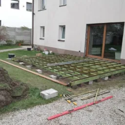 Konstrukcja pod taras w trakcie budowy: zielona rama z belek na betonowych bloczkach, narzędzia na żwirze, ziemia, dom z oknami w tle. Widok z boku.