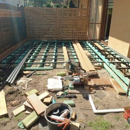 Łukasz Dziklinski - Montaż tarasu: widok na zieloną konstrukcję nośną, częściowo ułożone deski, narzędzia (piła) i materiały budowlane na ziemi. W tle drewniany płot i ściana budynku.