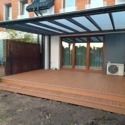 Łukasz Dziklinski - Zadaszony taras z kompozytowych desek w odcieniach brązu, z widoczną konstrukcją pergoli i fragmentem ogrodzenia. Na tarasie stoi klimatyzator.