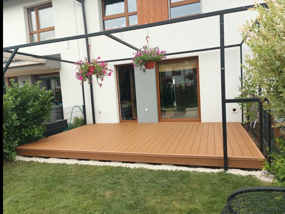 Nowoczesny taras kompozytowy z metalową konstrukcją pergoli, otoczony zielenią i białym żwirem, z doniczkami z kwiatami zwisającymi z ramy.