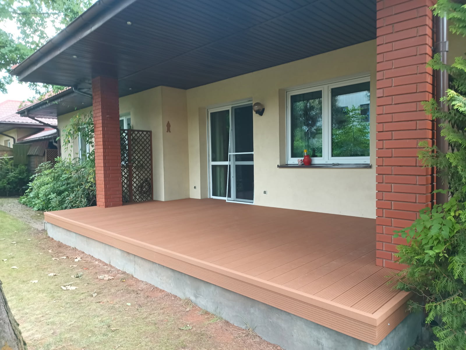 Nowoczesny taras kompozytowy w kolorze brązu przylegający do domu z ceglaną kolumną i dużymi oknami, otoczony zielenią. Widok z zewnątrz.