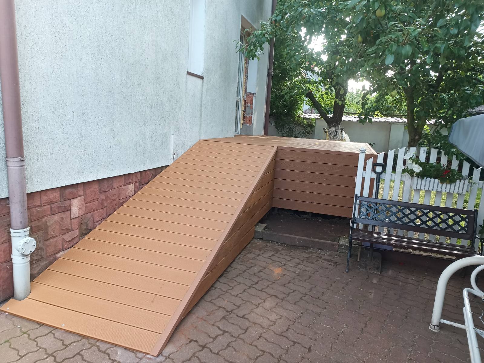 Rampa z desek kompozytowych prowadząca do podwyższonego tarasu przy budynku. Widoczna ławka i fragment ogrodu w tle.