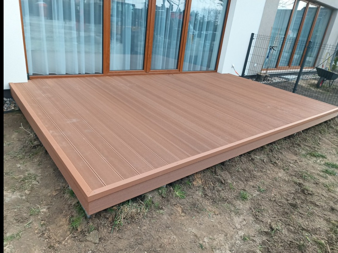 Nowy, brązowy taras kompozytowy przy białym domu z dużymi oknami. Widoczna ziemia wokół. Świeżo położone deski, brak mebli ogrodowych.