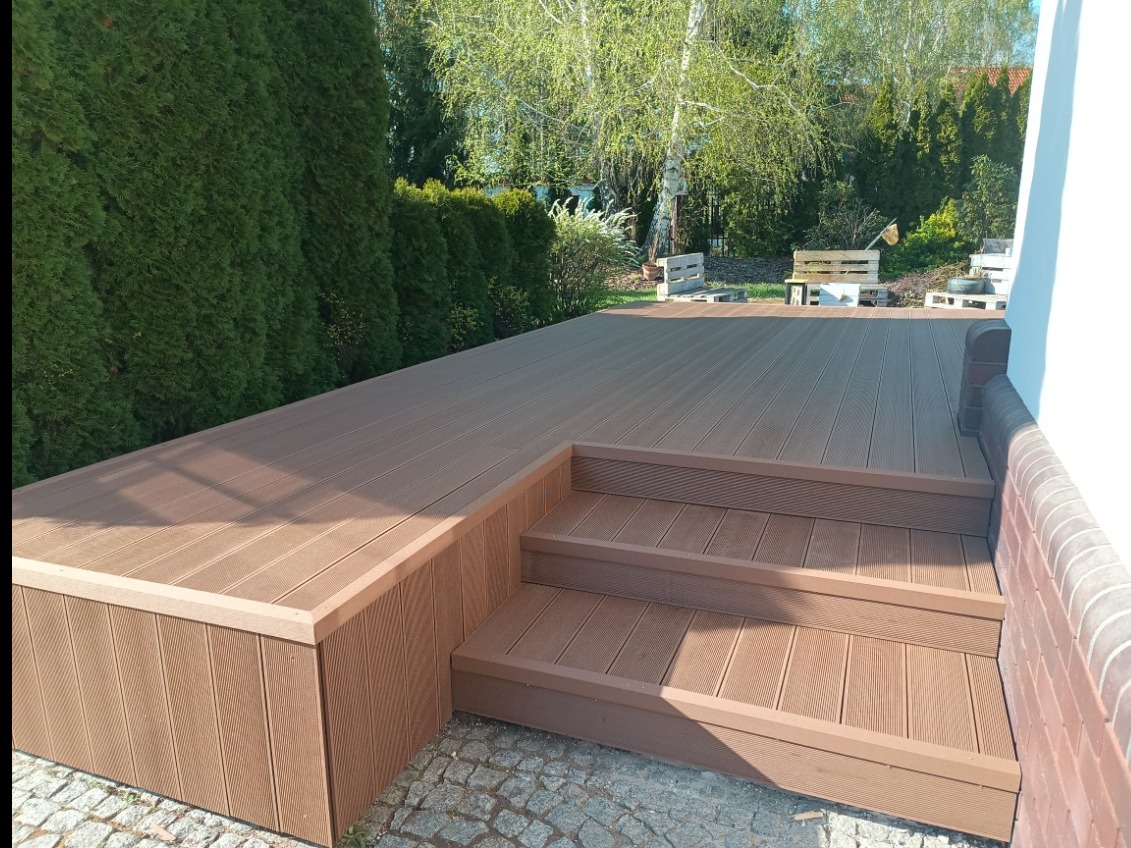 Nowoczesny taras kompozytowy z trzema schodkami, wykończony panelami w odcieniach brązu. W tle widoczne drzewa i krzewy, tworzące naturalne otoczenie.