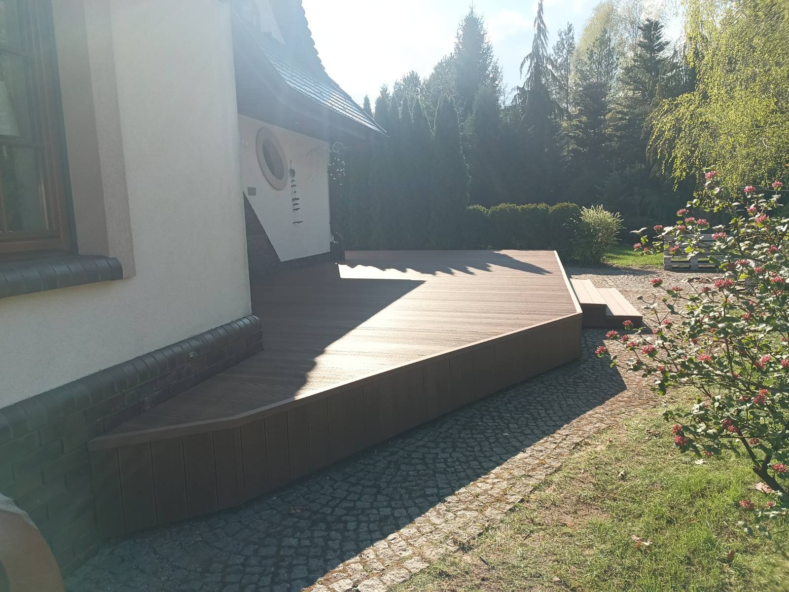 Nowoczesny taras z brązowych desek kompozytowych przylegający do domu, z widocznym fragmentem ogrodu i kostki brukowej. Słoneczny dzień.