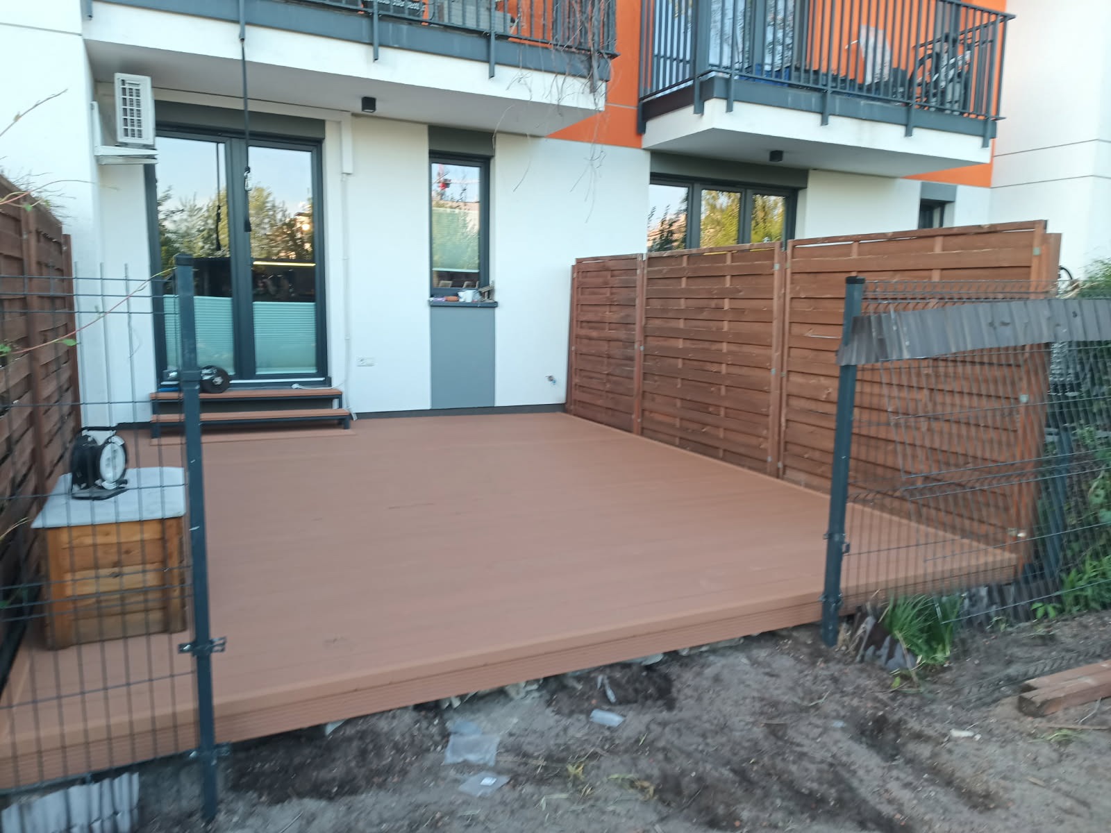 Nowoczesny taras kompozytowy przy budynku mieszkalnym, z drewnianym ogrodzeniem i metalową siatką. Widoczne elementy wykończeniowe i otoczenie.