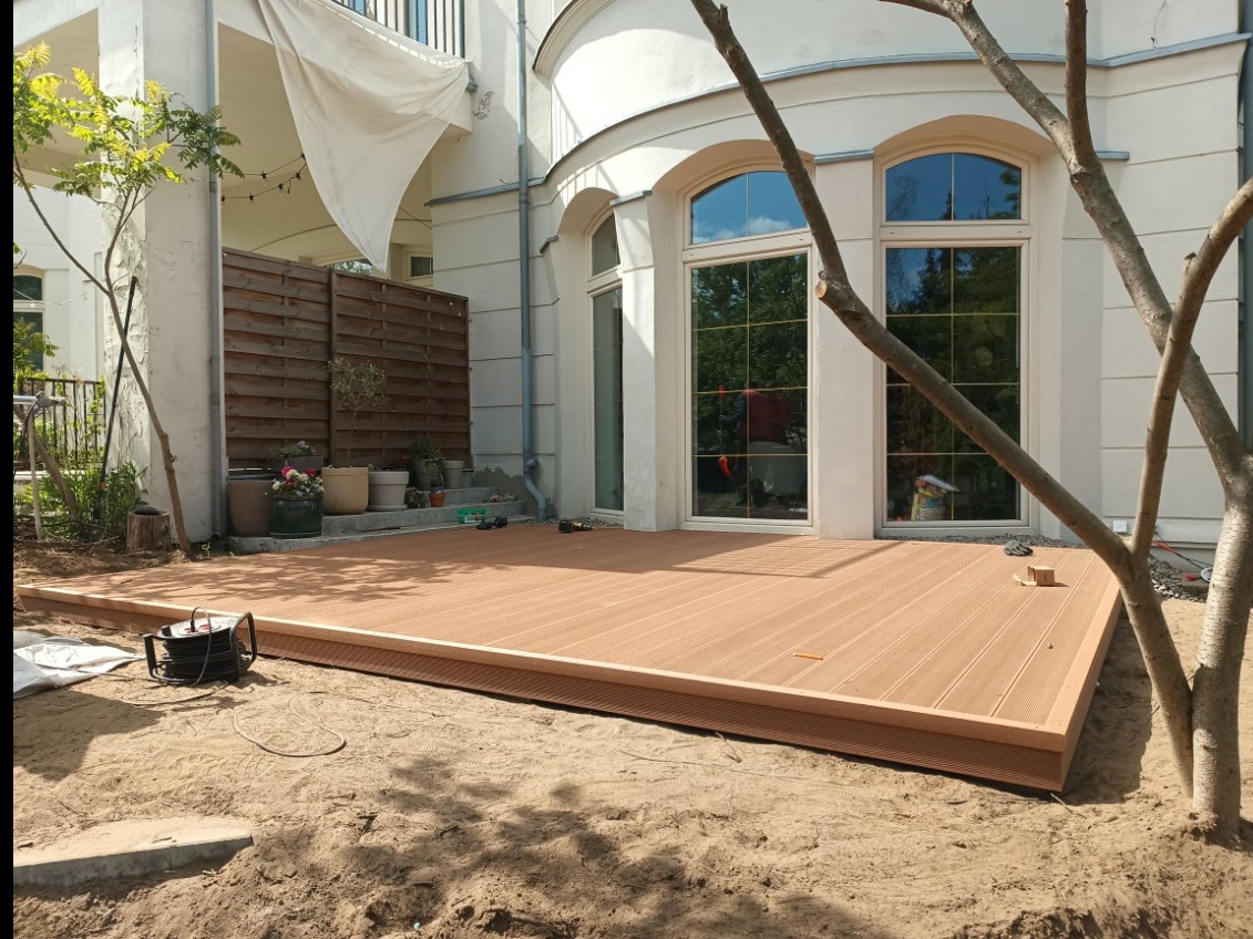 Nowoczesny taras z desek kompozytowych w odcieniach brązu, z widocznymi detalami montażu i elementami krajobrazu, takimi jak drzewo i jasna elewacja budynku.