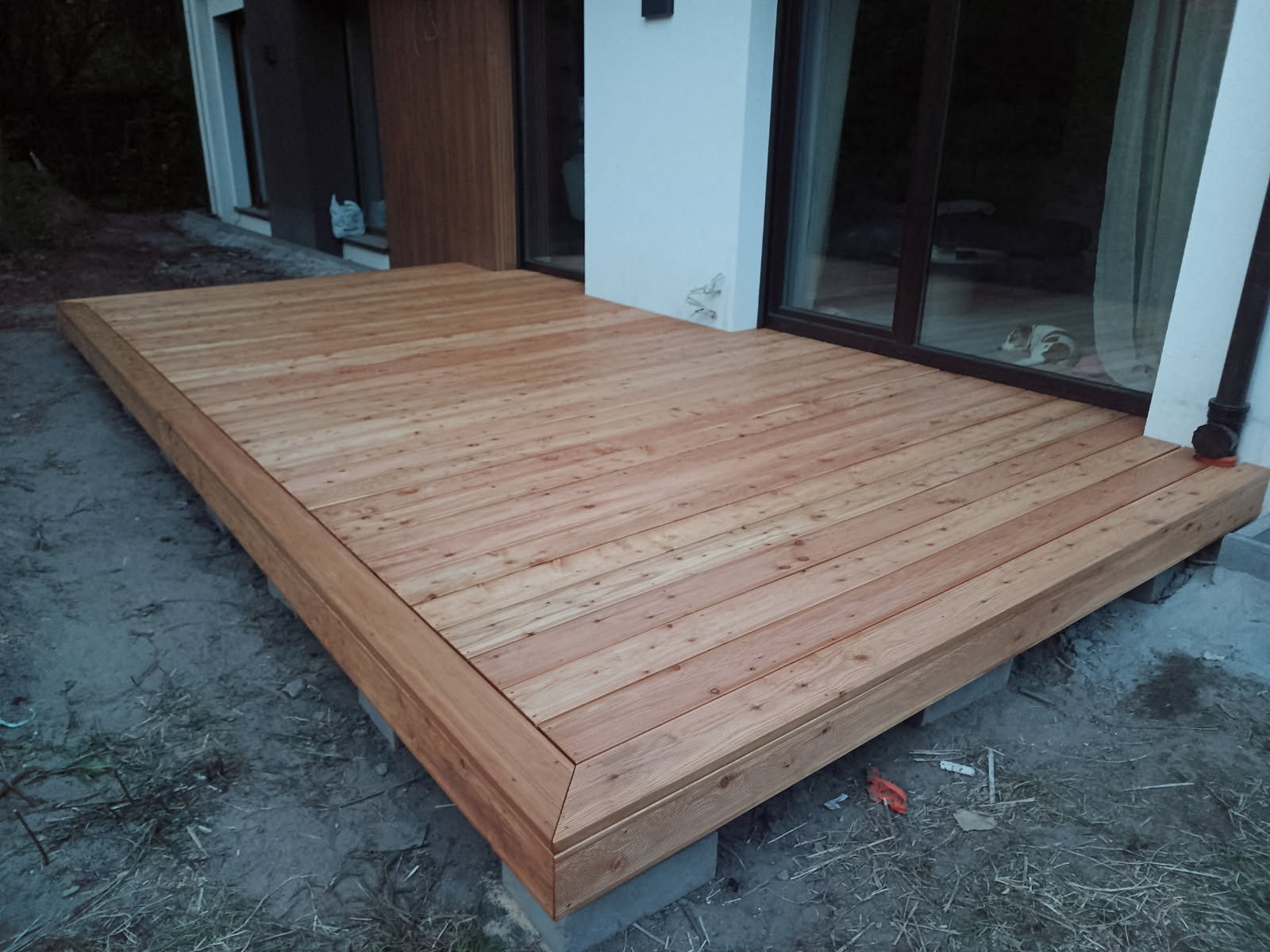 Nowoczesny taras drewniany z jasnego drewna, wykończony listwami, na betonowych bloczkach, przylegający do budynku z dużym oknem przesuwnym.