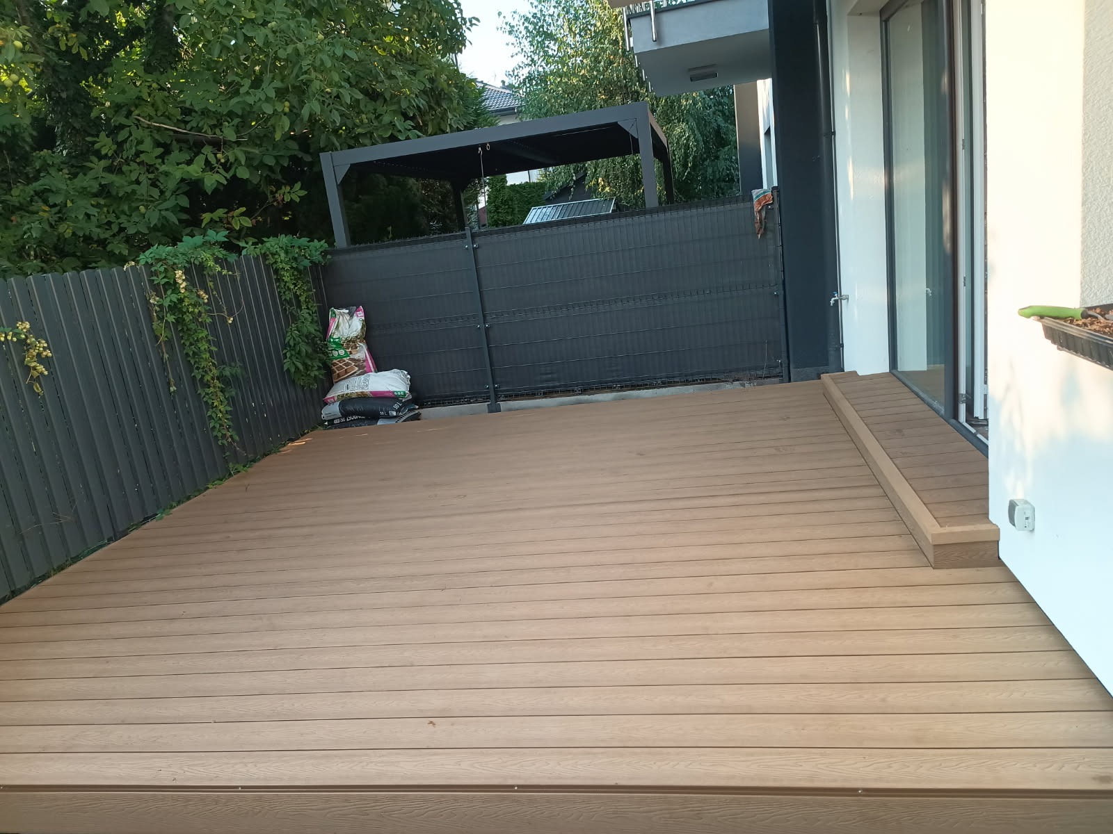 Nowoczesny taras z deski kompozytowej w odcieniach brązu, z widoczną pergolą i workami ziemi przy płocie. Widok na taras z niskiej perspektywy.