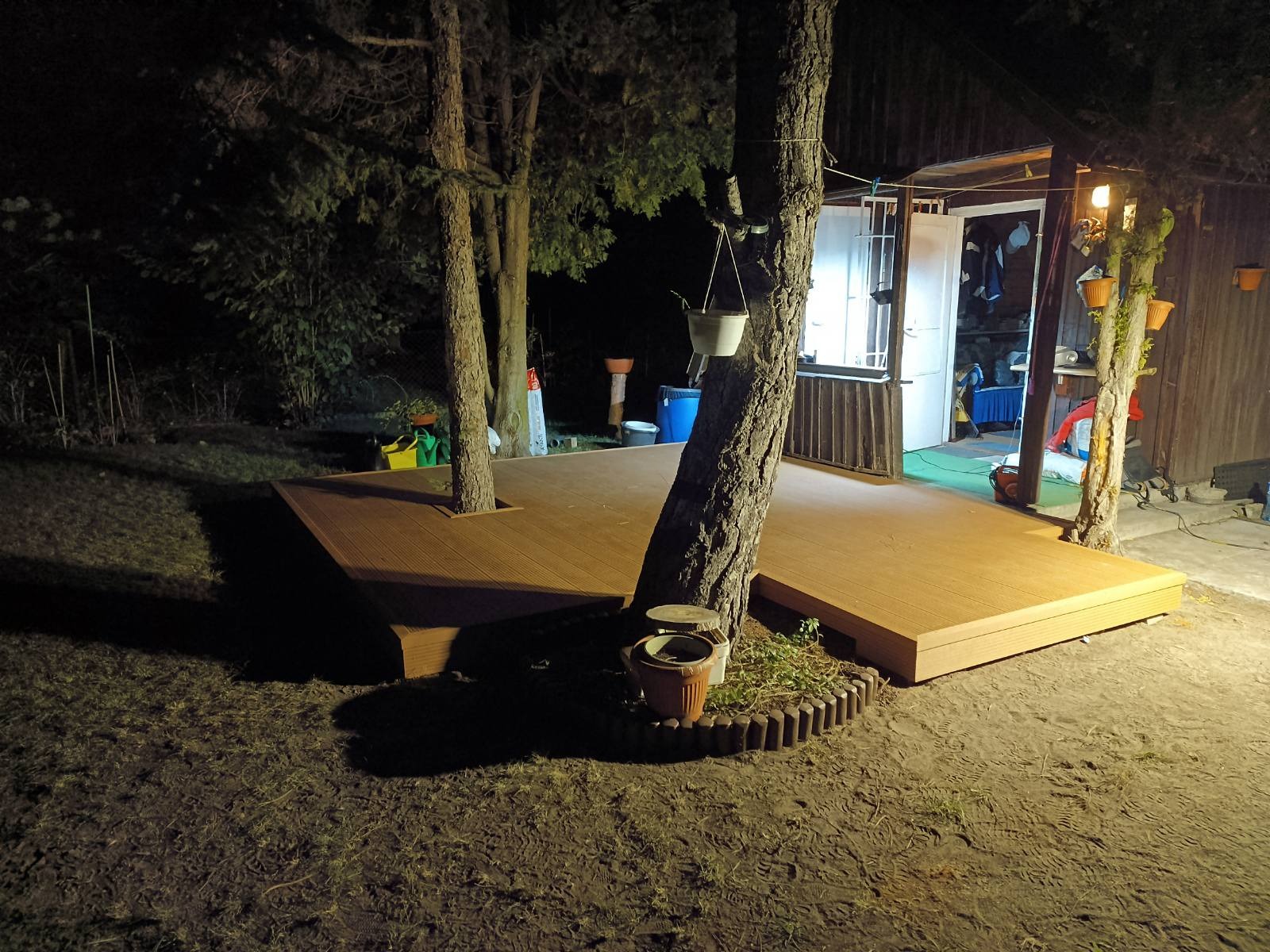 Nowoczesny taras kompozytowy z oświetleniem nocnym, zbudowany wokół drzew. Donice i elementy dekoracyjne dodają uroku przestrzeni.