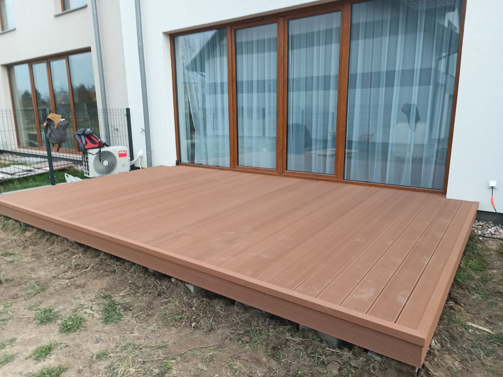 Nowoczesny taras kompozytowy w kolorze brązowym, przylegający do białego budynku z dużymi oknami. Widoczne detale konstrukcji i otoczenie z elementami zieleni.