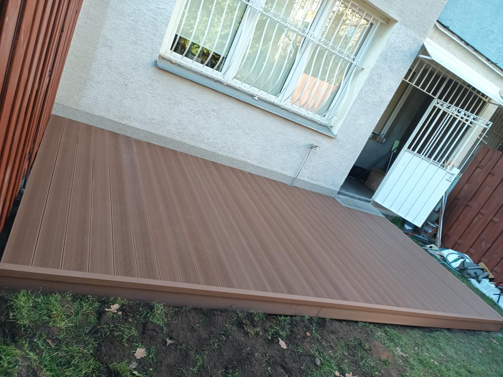 Nowy, brązowy taras kompozytowy przylegający do budynku z oknem w kratkę. Widoczne zielone trawniki i częściowo ogrodzenie. Ukończony montaż.