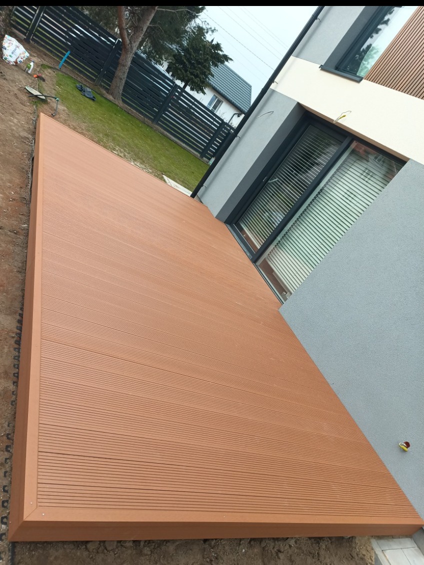 Nowy, brązowy taras kompozytowy przylegający do budynku z szarą elewacją. Widoczny fragment trawnika i ogrodzenia. Świeżo zakończone prace.