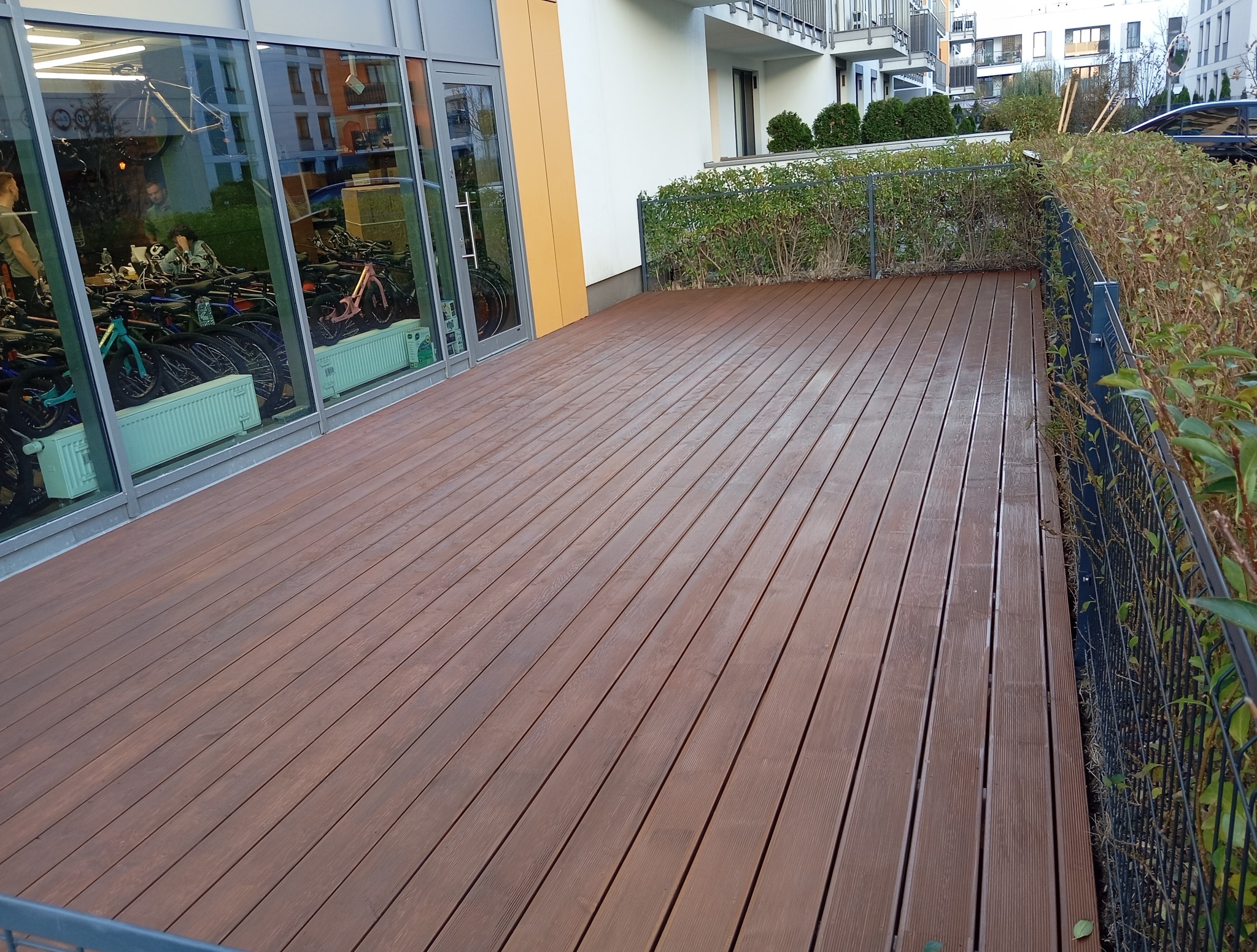 Nowoczesny taras z desek kompozytowych w odcieniach brązu, przylegający do budynku z dużymi oknami i zielenią w tle. Widoczna metalowa balustrada.