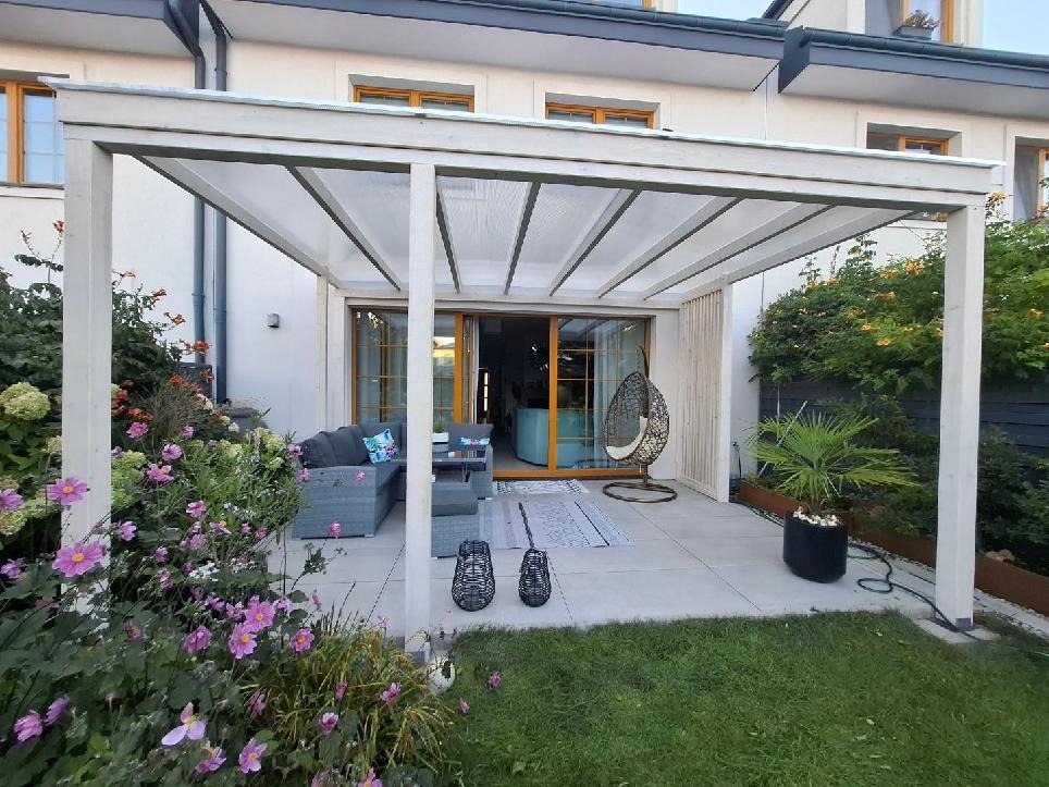 Biała pergola z przezroczystym zadaszeniem nad tarasem. Szara sofa, dywan i wiszące krzesło. Kwiaty i zieleń w ogrodzie. Dwie czarne latarnie.