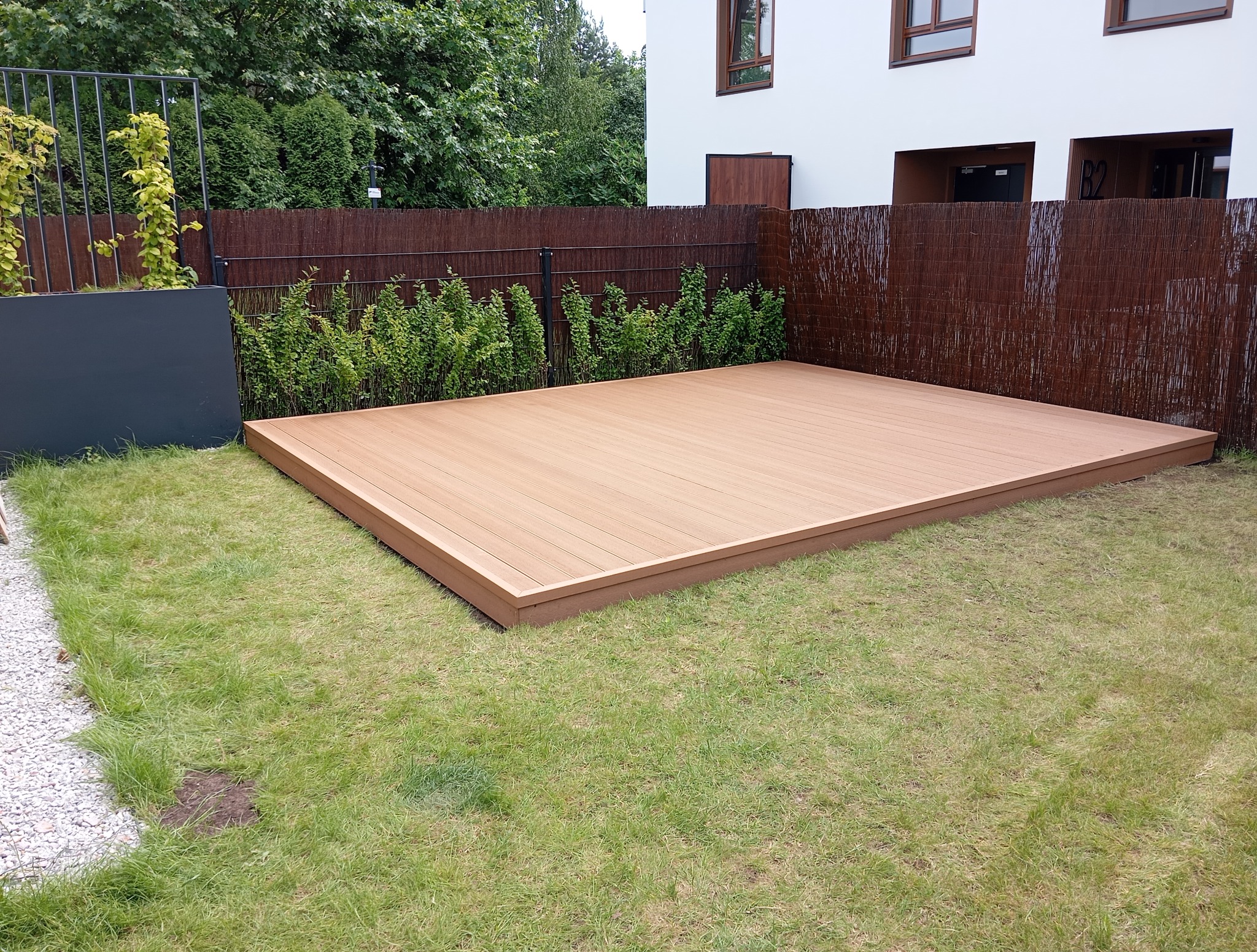 Nowoczesny taras kompozytowy w ogrodzie, z brązowym, plecionym ogrodzeniem w tle i białym budynkiem. Minimalistyczny design, trawa i zieleń wokół.