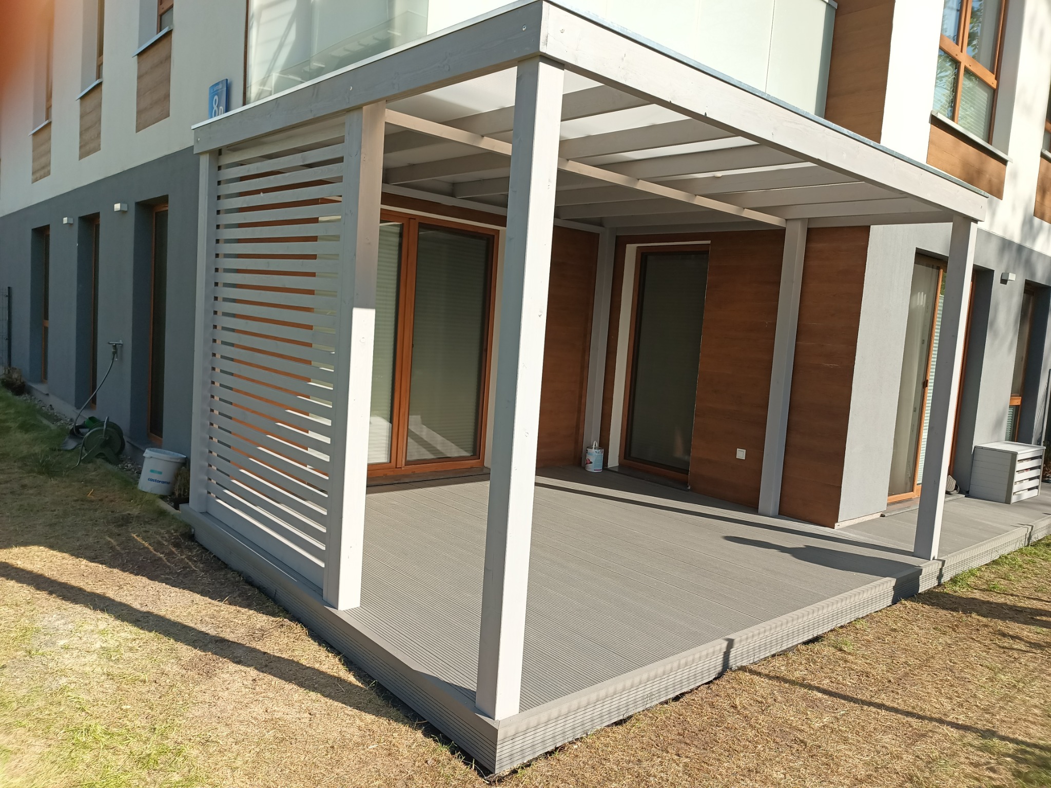 Nowoczesna pergola z szarego drewna i tarasem kompozytowym w Pruszkowie. Widoczne poziome lamele i elementy konstrukcyjne. Trawa dookoła tarasu.