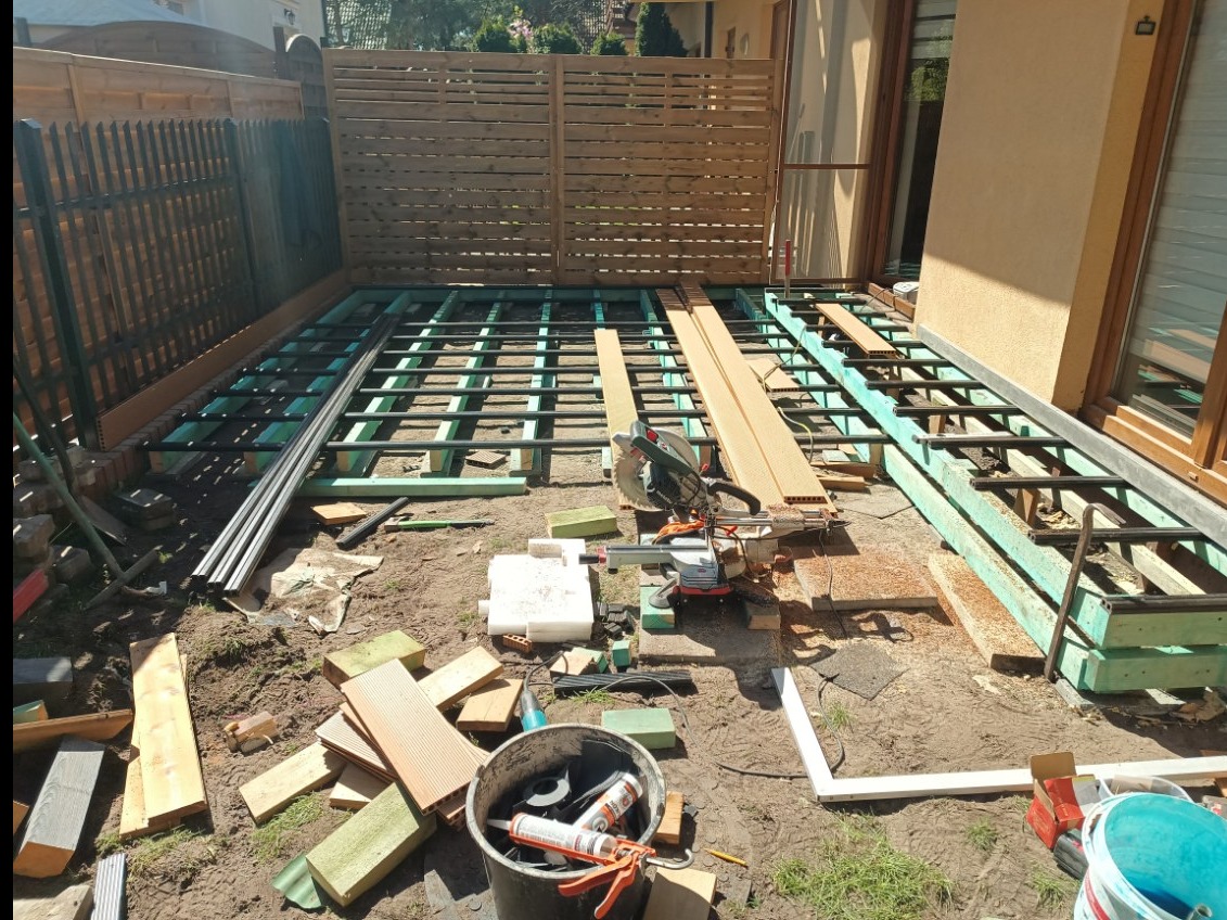 Montaż tarasu: widok na zieloną konstrukcję nośną, częściowo ułożone deski, narzędzia (piła) i materiały budowlane na ziemi. W tle drewniany płot i ściana budynku.