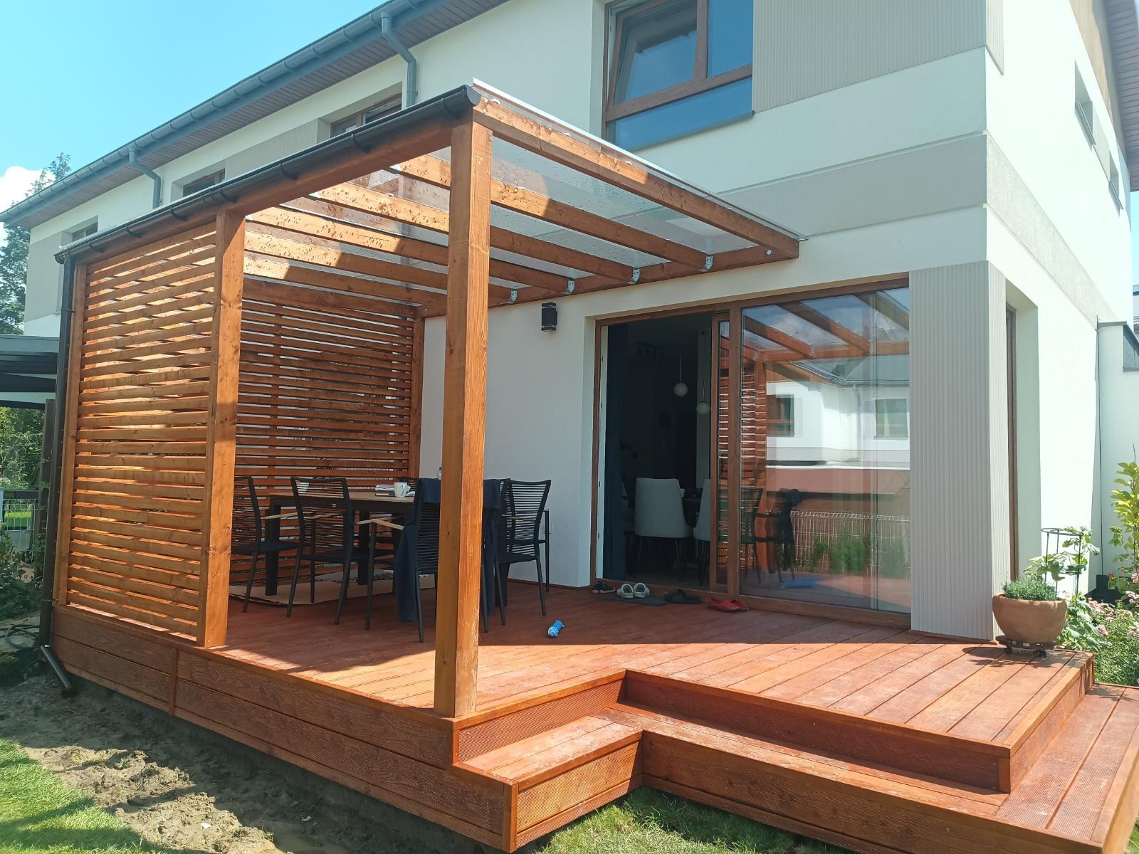 Drewniany taras z pergolą i zadaszeniem z poliwęglanu przy domu. Widoczne schody, stół z krzesłami i ażurowa ścianka. Słoneczny dzień.
