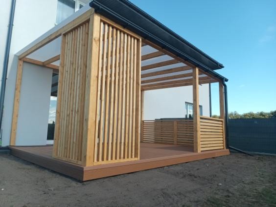 Drewniana pergola z zadaszeniem z poliwęglanu, z elementami ażurowymi, na brązowym tarasie kompozytowym, przylegająca do białego budynku. Błękitne niebo w tle.