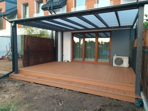 Zadaszony taras z kompozytowych desek w odcieniach brązu, z widoczną konstrukcją pergoli i fragmentem ogrodzenia. Na tarasie stoi klimatyzator.