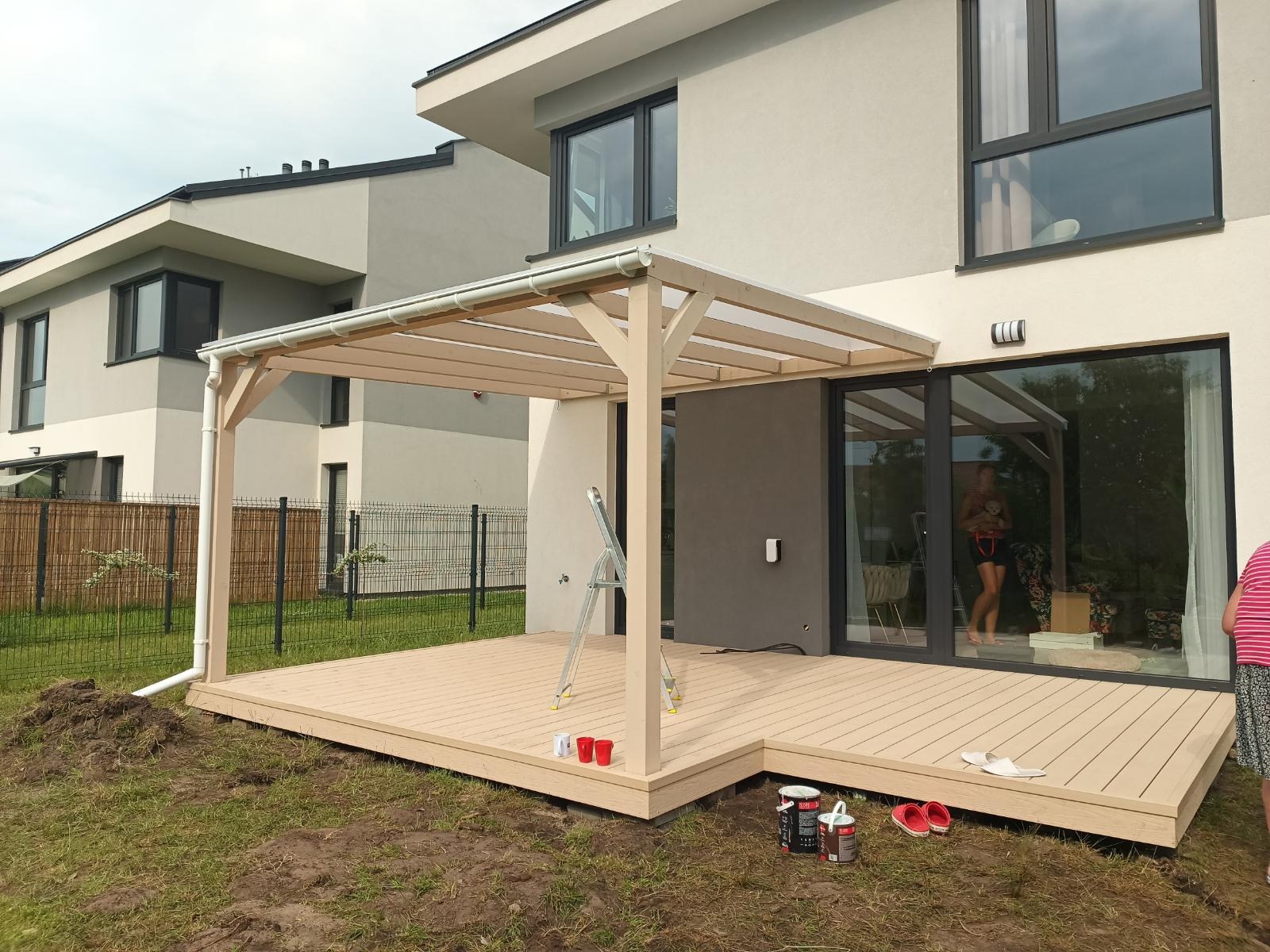 Jasny taras z zadaszeniem przy nowoczesnym domu. Widoczne farby, drabina i elementy wykończeniowe. Częściowo wykończony teren wokół tarasu.