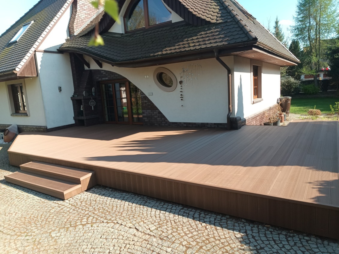 Nowoczesny taras kompozytowy z brązowych desek, z niskimi schodkami, przylegający do domu z oryginalną architekturą i okrągłym oknem. Kostka brukowa na pierwszym planie.