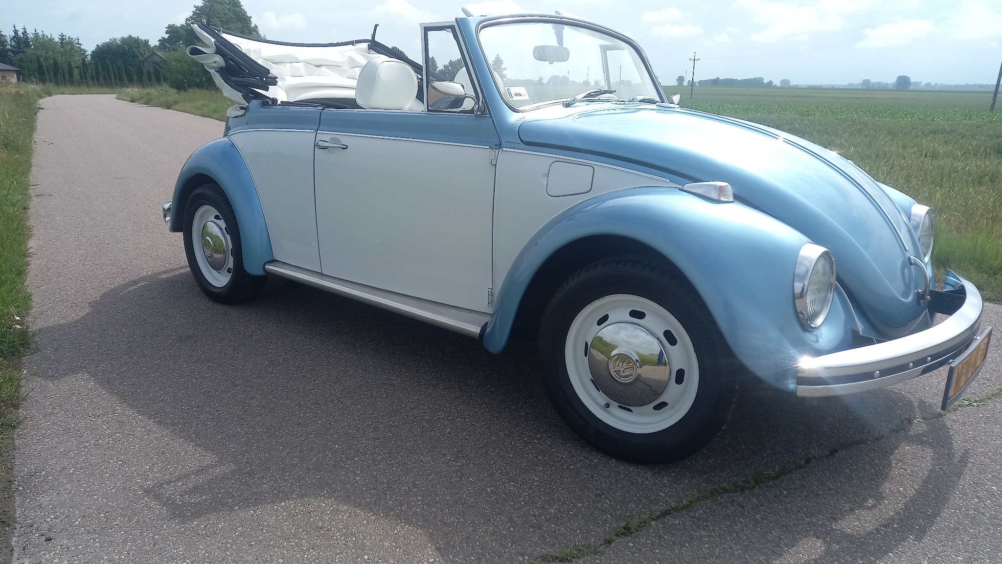 Błękitno-biały, odrestaurowany Volkswagen Garbus Cabrio na asfaltowej drodze w słoneczny dzień. Klasyczny model z otwartym dachem i polerowanymi felgami.