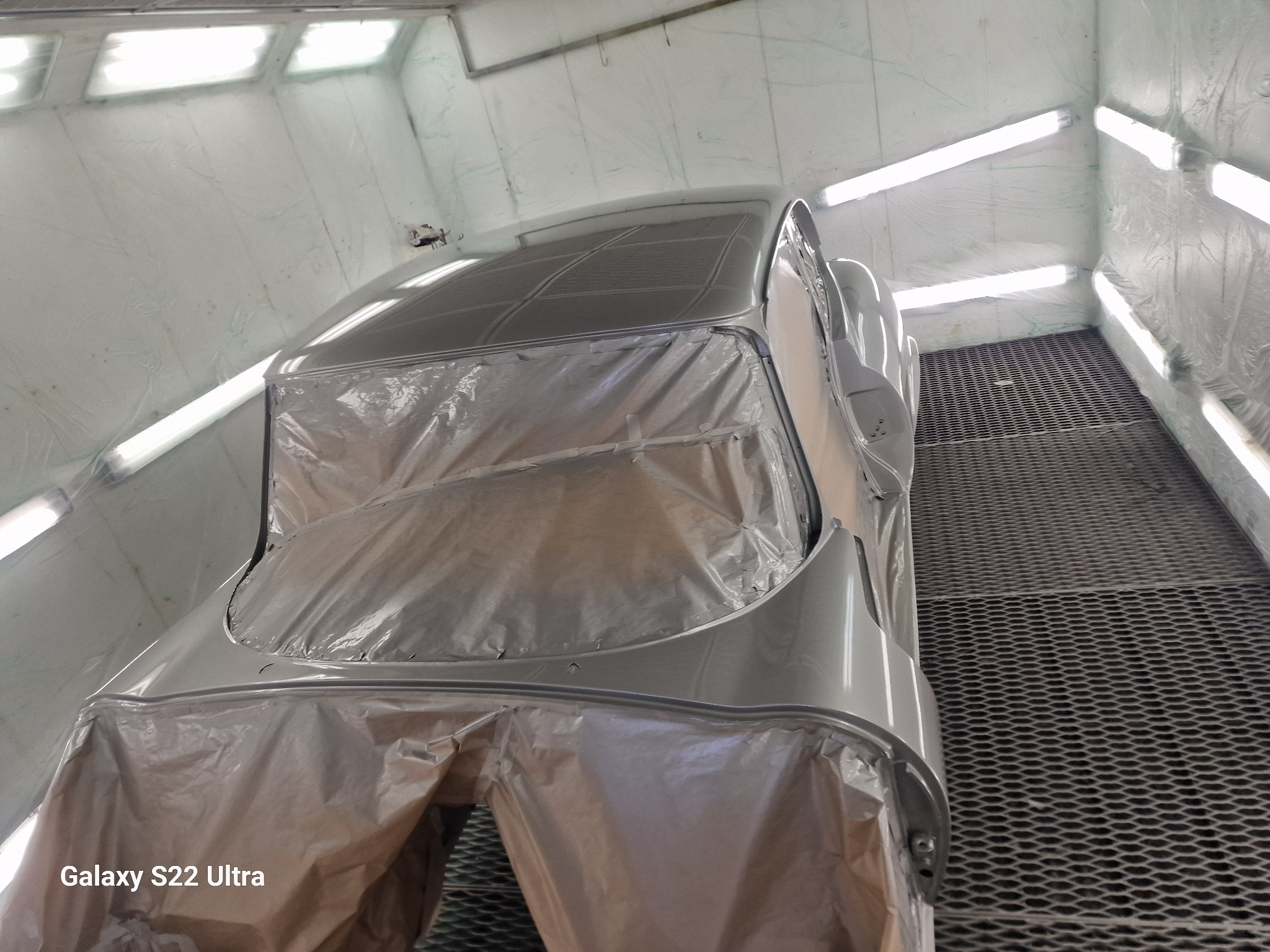 Klasyczny samochód w trakcie lakierowania, częściowo zakryty folią ochronną, w jasnym pomieszczeniu lakierniczym z kratą podłogową.