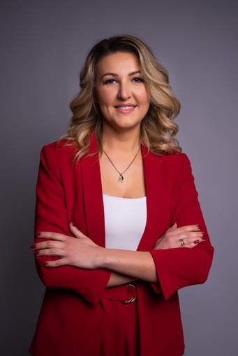 Portret kobiety w czerwonym żakiecie, z blond włosami i uśmiechem, na szarym tle. Skrzyżowane ręce dodają pewności.