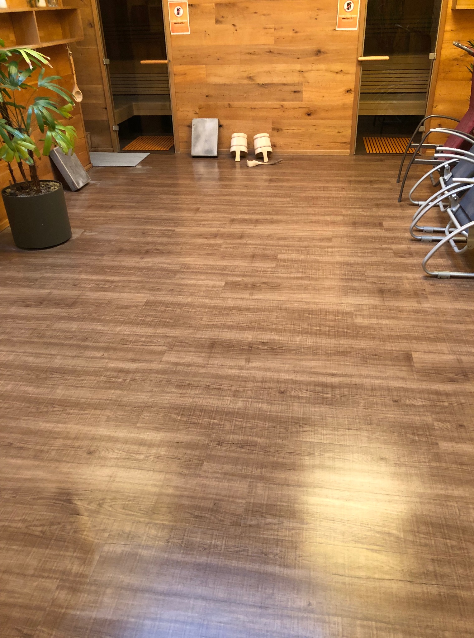 Drewniana podłoga w saunie z widocznymi wejściami do kabin, leżakami i rośliną w doniczce. Para klapek i kamień leżą na podłodze.