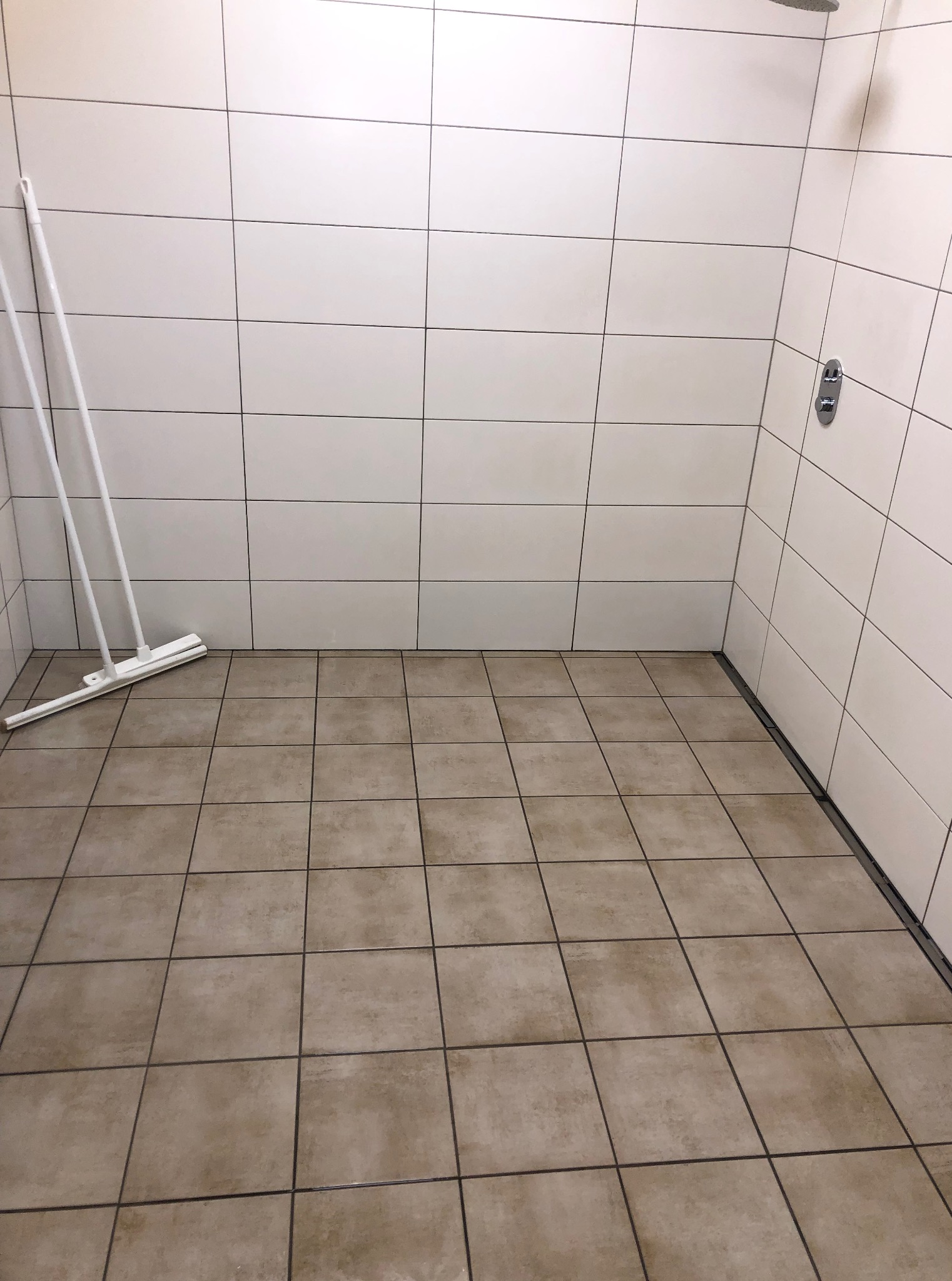 Umyta kabina prysznicowa z beżowymi płytkami podłogowymi i białymi ściennymi. W rogu stoi biała ściągaczka do wody.