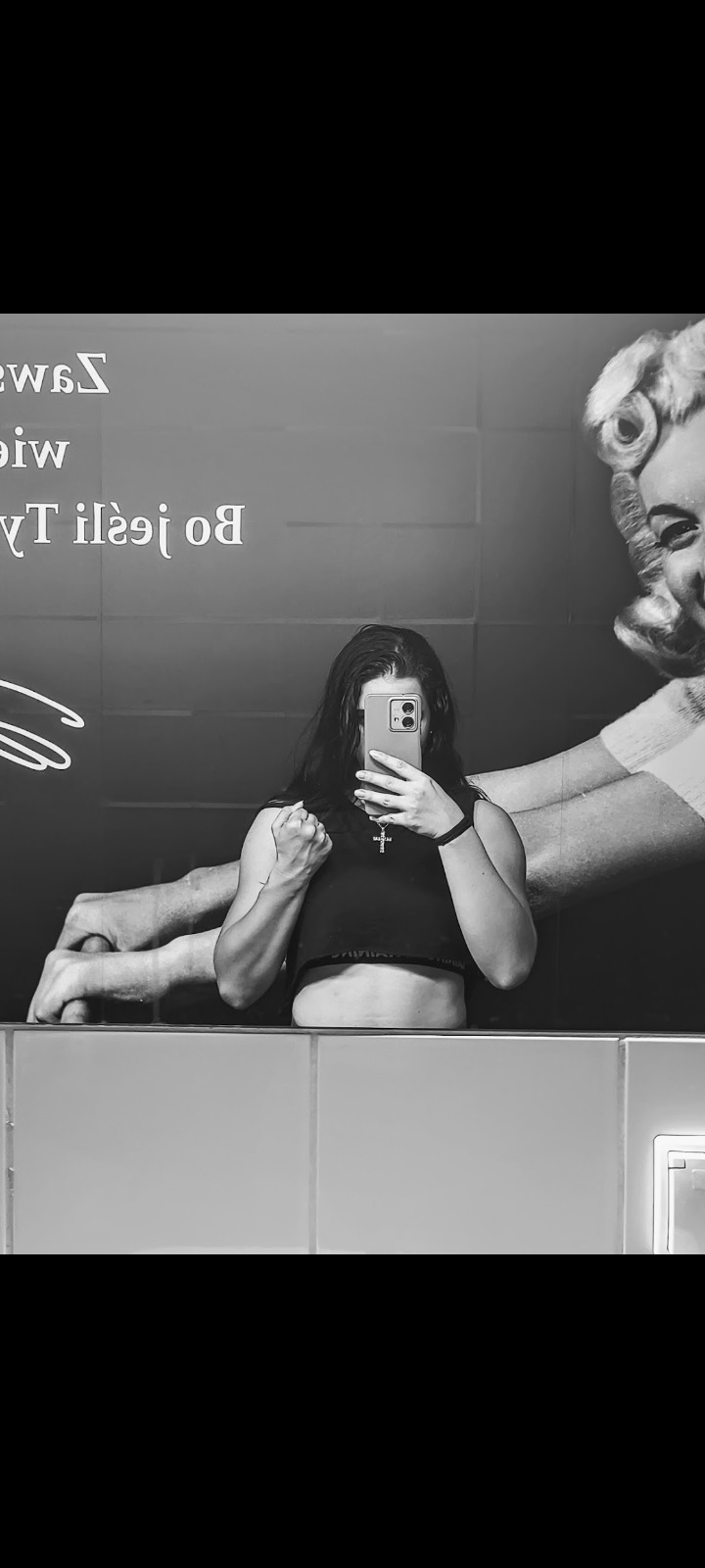 Kobieta w sportowym stroju robi selfie w lustrze łazienki z artystycznym nadrukiem, prezentująca sylwetkę po treningu. Widoczny krzyżyk na szyi i zegarek sportowy.