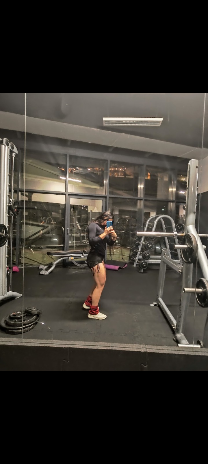 Kobieta w sportowym stroju, z obciążnikami na kostkach, robi selfie w lustrze na siłowni. W tle widoczne maszyny do ćwiczeń i okno.
